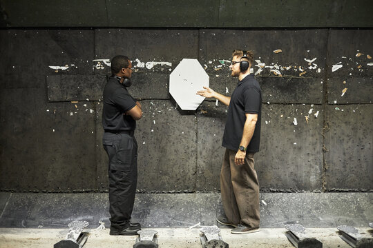 Caucasian man explaining shooting results to Black male customer at indoor shooting range, both wearing protective ear muffs and standing near paper target