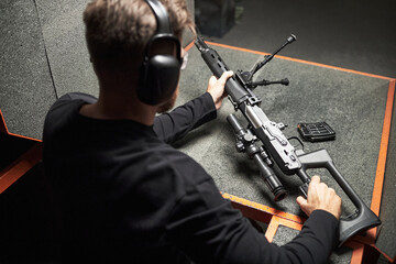 Rear view of man wearing protective earmuffs sitting at shooting range table holding sniper rifle preparing for target practice with ammunition magazine