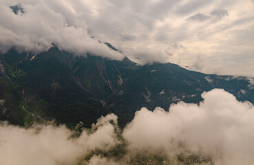 Mountain peaks covered in snow and clouds with green forest trees in foreground, dramatic sky