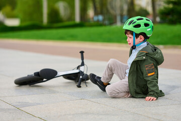 child with green helmet sitting beside fallen bicycle on urban path. young boy's learning moment in...