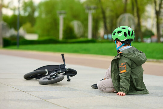 child sitting beside fallen bicycle, wearing green helmet and jacket, looks away in park setting. represents outdoor adventure, exploration, childhood resilience, and learning moments