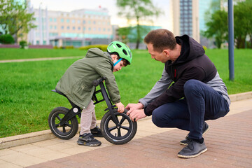father helping child with balance bike in urban park, encouraging outdoor play. kid wears helmet...
