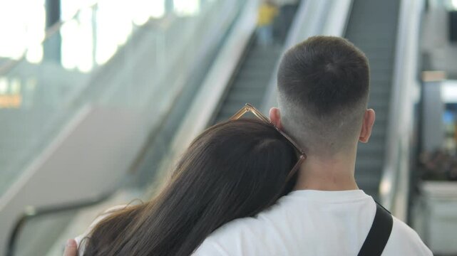 Man and woman are hugging in an airport. The woman has long hair and the man has a short haircut. They are both wearing white shirts