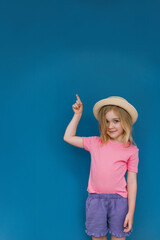 Young girl in pink shirt and hat points against vibrant blue wall background