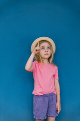 Young girl with a straw hat pointing upwards against a blue wall in bright summer attire
