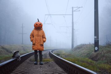 Halloween girl in pumpkin mask with scarecrow severed head poses on abandoned train tracks in thick autumn fog. Creepy railway moment with mysterious mood and fall holiday style