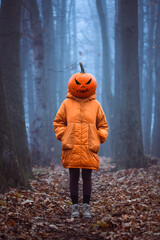 Girl in orange jacket with a spooky carved pumpkin head on Halloween in the misty autumn forest. Fall holiday concept