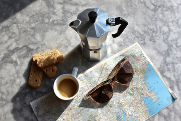 Flat lay of a travel scene with a moka pot, espresso cup, cookies, sunglasses, and a city map arranged on a marble surface