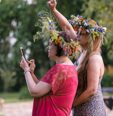 Two women with summer solstice wreaths on their heads stand side by side and look at the phone held...