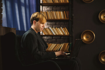 Man working on laptop in a dark, atmospheric room. Sitting in a chair, focused, with bookshelves...