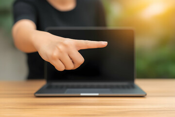 A person points at a closed laptop sitting on a wooden table with a blurred natural background and warm sunlight.