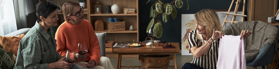 Two middle aged Caucasian women sitting on sofa, holding wine glasses watching middle aged Caucasian woman showing clothing item in cozy living room setting during friendly gathering