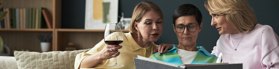 Three middle aged Caucasian women sitting together on sofa, one holding wine glass, all looking at photo album and engaging in conversation, casual indoor setting