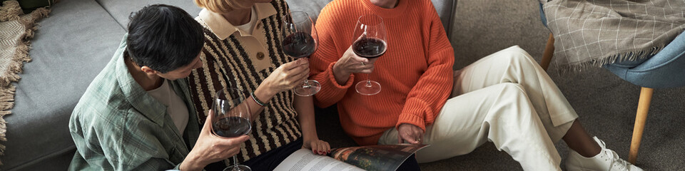 Three young adult women sitting on floor holding wine glasses, reading magazine together, relaxing in casual setting, two with short hair, one with medium length hair, all appearing Caucasian