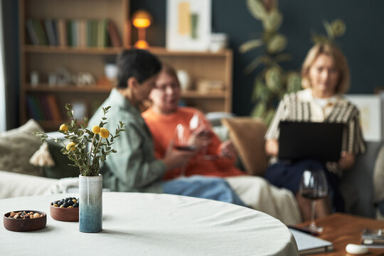 Three Caucasian middle aged women sitting together in living room, two women holding wine glasses and talking while third woman using laptop in background, blurred focus on group