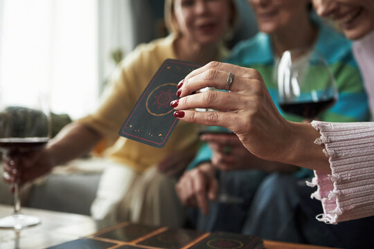 Group of young adult Caucasian women playing card game together, holding cards and drinking red wine, close up of hand with ring in foreground, friends enjoying leisure activity indoors
