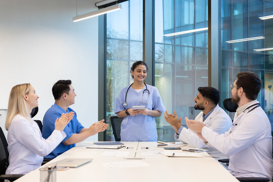 A diverse medical team applauds in a modern office setting. A female doctor stands, holding a tablet and smiling.