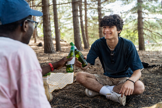 Two happy diverse young friends toasting with beer bottles at a campsite in the forest