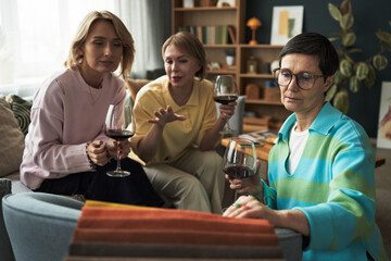 Three Caucasian middle aged women sitting together holding wine glasses, engaging in conversation in living room setting, one woman gesturing while others listening attentively