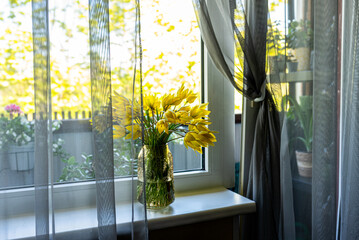 A windowsill with a vase of yellow tulips, softly lit by warm natural light. The balcony door is...