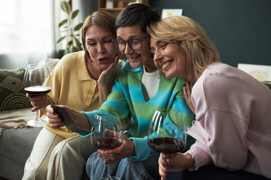 Three middle aged Caucasian women sitting close together on sofa smiling and looking at smartphone while holding wine glasses, enjoying friendly moment during adult girlfriends gathering - Powered by Adobe
