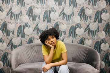 Bored young woman with afro hair is sitting on a sofa resting her head on her hand, showing boredom...