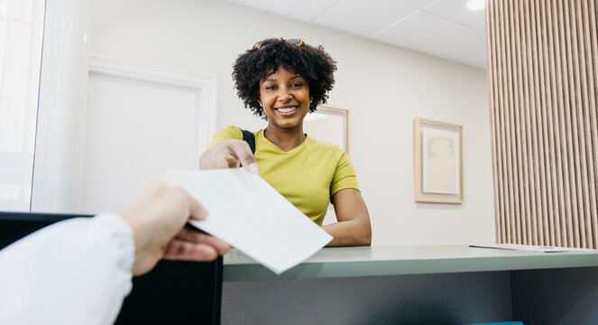 Smiling patient handing paperwork from the receptionist, scheduling appointment in modern clinic