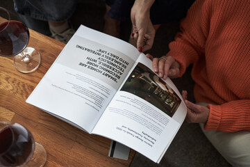 Naklejka premium Two young adult Caucasian women sitting together reading open magazine on wooden table, hands visible holding pages, glasses of red wine nearby