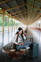 Happy young caucasian woman with phone and headphones stands on station platform. Girl blonde smiles looking aside, wears jacket. Technology concept, lifestyle