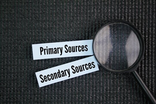 A magnifying glass and colored paper with the words Primary and Secondary Sources. Primary sources provide direct, firsthand accounts of events or topics, while secondary sources offer analysis, inter