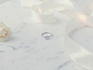 A sparkling diamond engagement ring in a silver setting is elegantly displayed on a white marble surface, accompanied by a soft white rose and ribbon.
