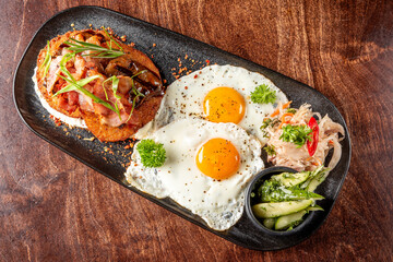 Breakfast plate with sunny-side-up eggs, crispy bacon, hash browns, pickled vegetables, and cucumber salad, served on a black plate on a wooden table