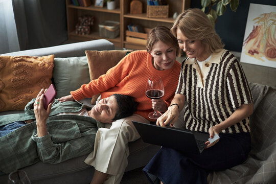 Three Caucasian young adult women relaxing on sofa, one lying down holding smartphone, two sitting close together holding wine glasses, all looking at laptop screen and smiling