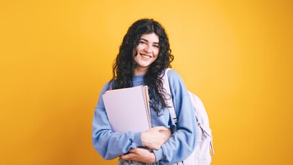 Smiling student holding books and wearing backpack on yellow background