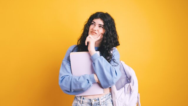 Pensive student thinking with backpack and notebooks on yellow background