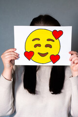 Woman holding smiling emoji mask with three hearts in front of face. Social media trends, Gen Z...