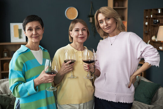 Three middle aged Caucasian women standing together holding wine glasses, smiling at camera, showing friendship and connection in casual indoor setting, celebrating adult girlfriends
