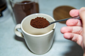 A hand holding a spoonful of ground coffee above a ceramic mug. Close-up