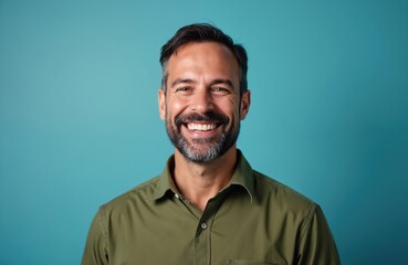 Fototapeta premium Happy middle-aged man smiles at camera against blue background. Portrait of cheerful 40s male with beard. Smiling adult person shows confidence. Smiling businessman in casual shirt. Positive emotions.