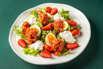 Fresh salad with mixed greens, boiled eggs with tomato relish, cottage cheese, cherry tomatoes, and strawberries on a white plate over a green background. Healthy and colorful dish.