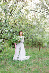 A young woman in a white dress stands in a blooming forest holding a bouquet of flowers. Light...