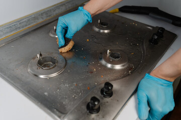 Housekeeper wearing blue gloves scrubbing dirty gas stove with sponge, removing grease and food...