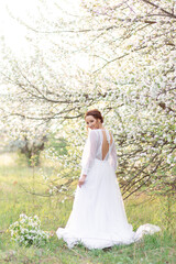 Fototapeta premium A young woman in a white dress stands against a background of flowering trees, surrounded by greenery and light, creating an atmosphere of spring and tenderness.