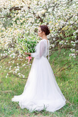 Bride in white dress stands among blossoming trees holding bouquet. Spring garden, delicate...