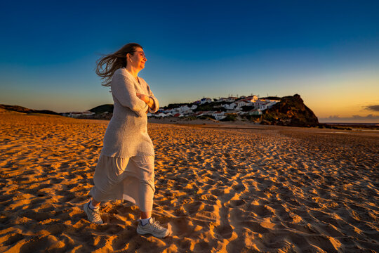Beautiful middle-aged woman walking on sandy beach on sunset on windy spring day. Side view. Monte Clerigo beach on Algarve coast in Portugal - Powered by Adobe