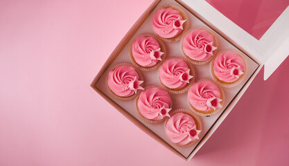 Flat lay of beautiful cupcakes with pink whipped cream cheese topping. A box of birthday cupcakes on a pink pastel background. Festive dessert delivery concept