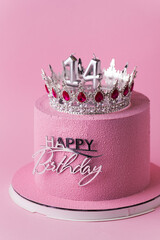 Beautiful luxury birthday anniversary cake for a girl with pink chocolate velvet coating decorated with silver crown and happy birthday topper. Pink background. Happy 14 years old celebration concept
