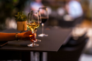 Close-up of a hand holding a glass of white wine at a stylish table, with blurred background and evening light, capturing a relaxed and elegant social moment.