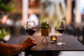 Close-up of a man's hand holding a glass of red wine at a stylish outdoor table, with candlelight and soft background, evoking a calm and refined evening mood.