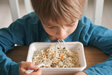 6 years old boy eats instant noodle from box. Child is eating spaghetti. Lunch time. A good appetite. Tasty children food. Close-up portrait. Carbs dinner. Fast food. Kid eating by himself with fork.